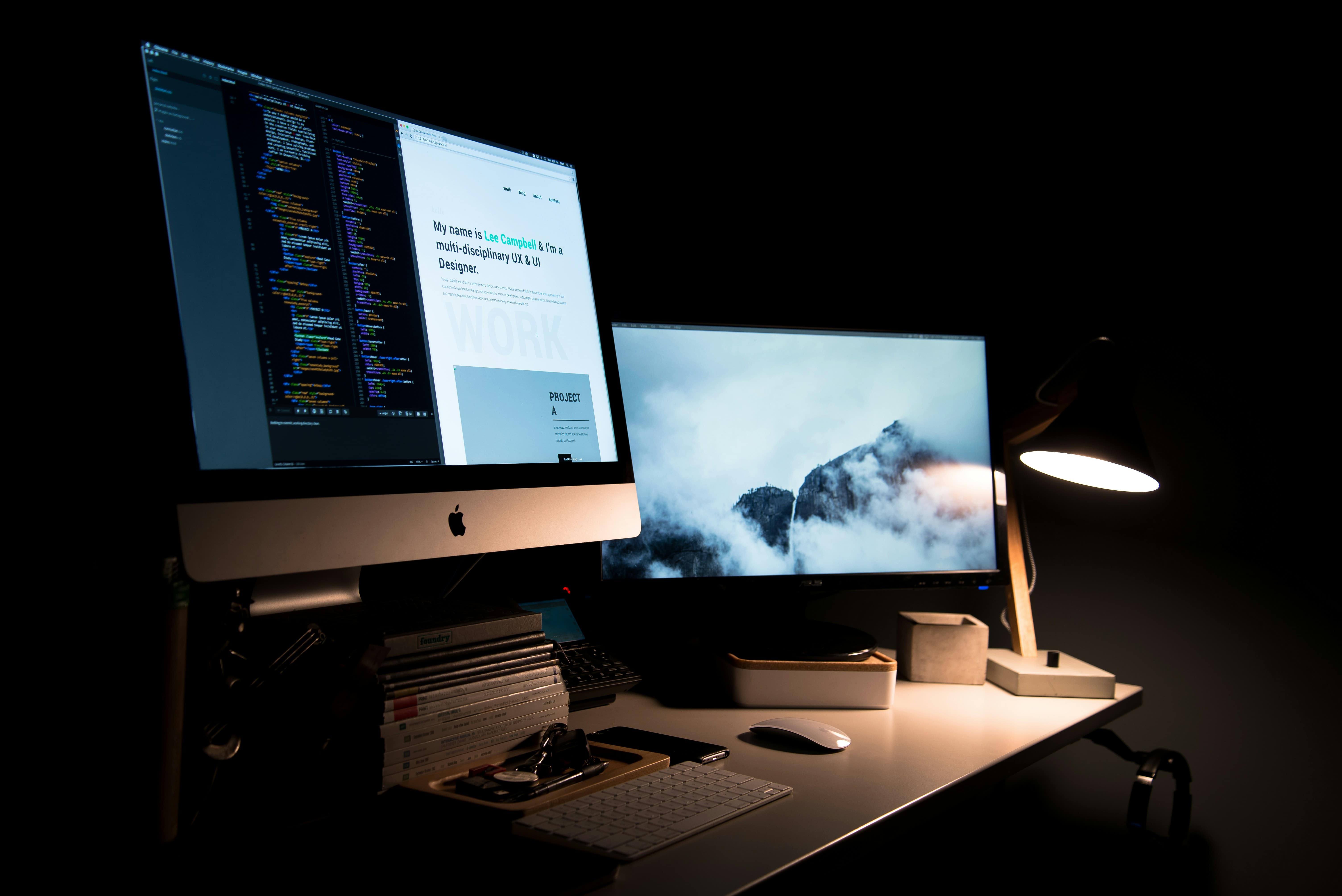 Two computer screens in the dim light of an office, one with code and a web page, the other with a mountain landscape