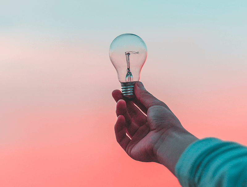 A hand holding out a clear light bulb against a background fading from blue to pink